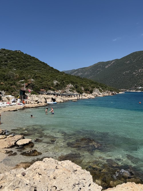 Swimmers wading in clear turquoise water by a rocky shore and a small pier, İnceboğaz Çınar Beach, Turkiye