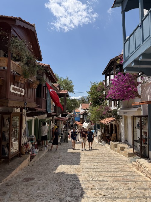 Pedestrians on a cobbled shopping street lined with wooden balconies, bougainvillea and a Turkish flag, Queen, Turkiye