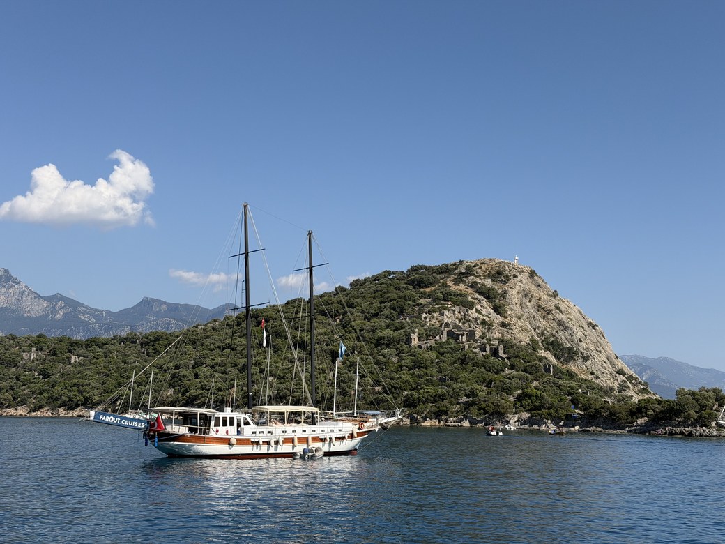 Two-masted gulet anchored in calm water below a forested hill with stone ruins, Santa Claus Island, Turkiye