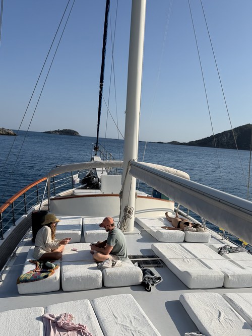 Three travelers lounging on white sun cushions on a gulet's foredeck, calm blue sea beyond, Santa Claus Island, Turkiye