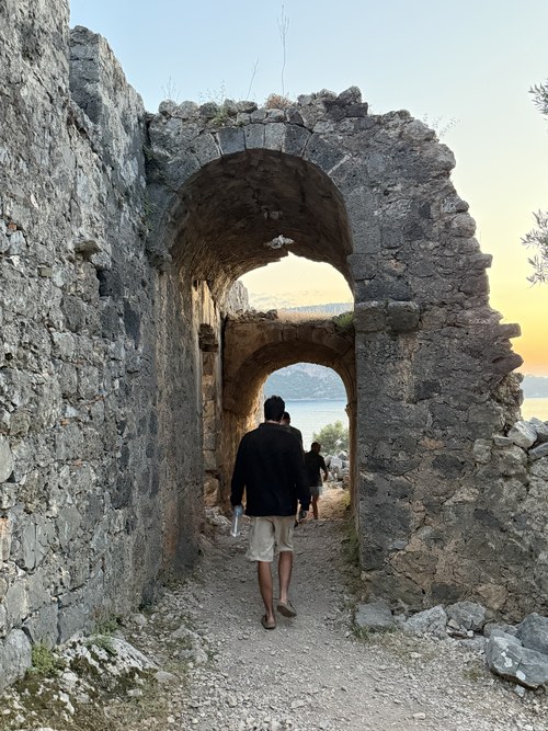Travelers walking through a ruined stone archway toward the sea at sunset, Santa Claus Island, Turkiye