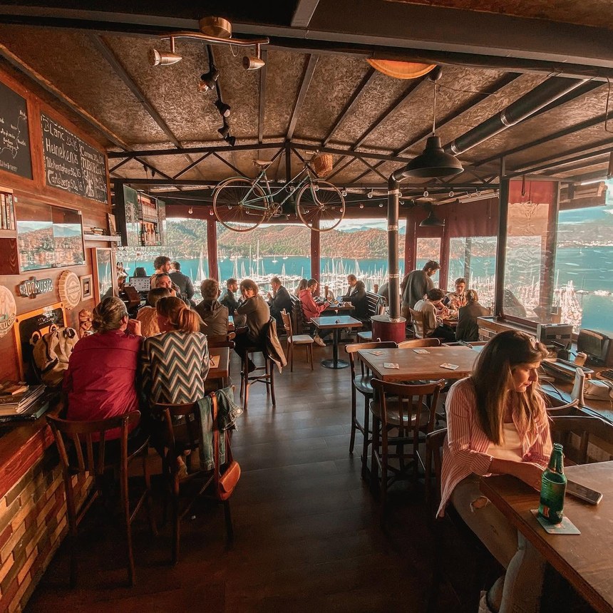 Crowded wooden bar with a vintage bicycle hung from the ceiling and the harbor and hills through the open windows, El Camino Pub & Hostel, Fethiye, Turkiye