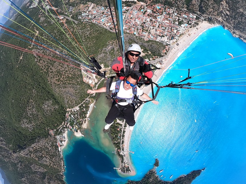 Selfie of a tandem paragliding pair above a turquoise lagoon, beach and hillside town, Fethiye Oludeniz, Turkiye