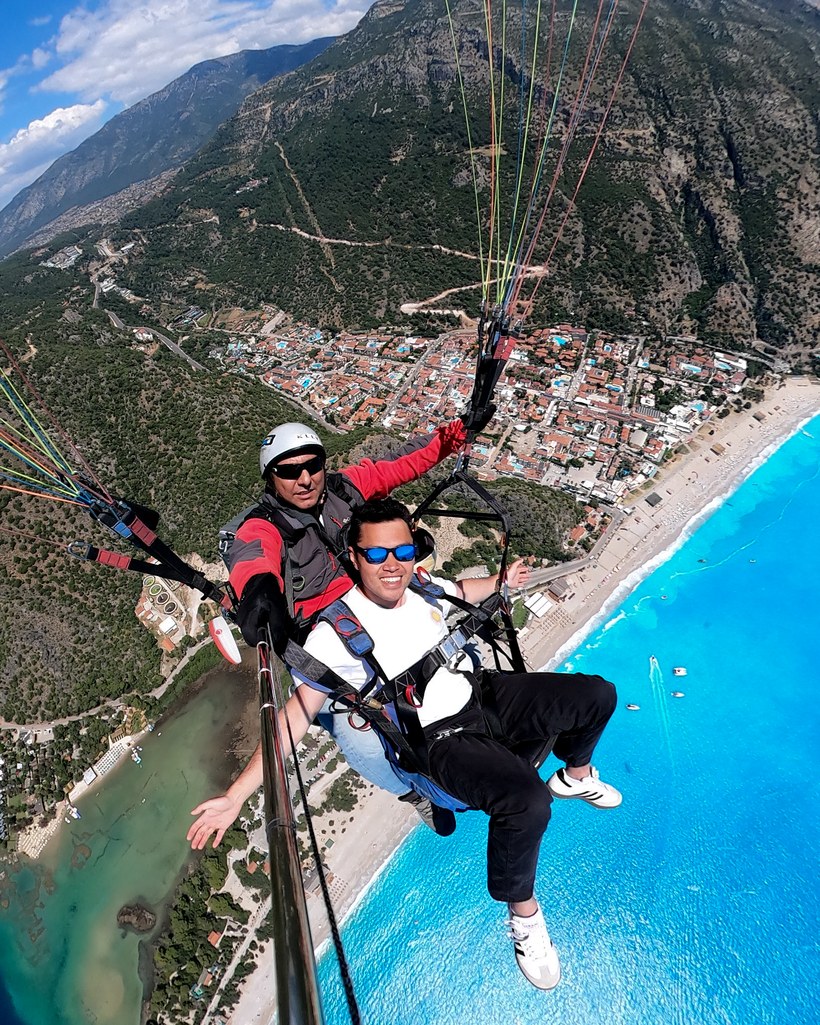 Two people paragliding in tandem over a turquoise sea, sandy beach and hillside town, Fethiye Oludeniz, Turkiye