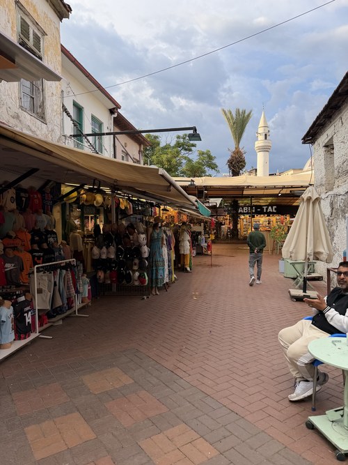 Brick-paved lane lined with clothing and hat stalls, a seated man and a minaret in the distance, Fethiye Old Town, Turkiye