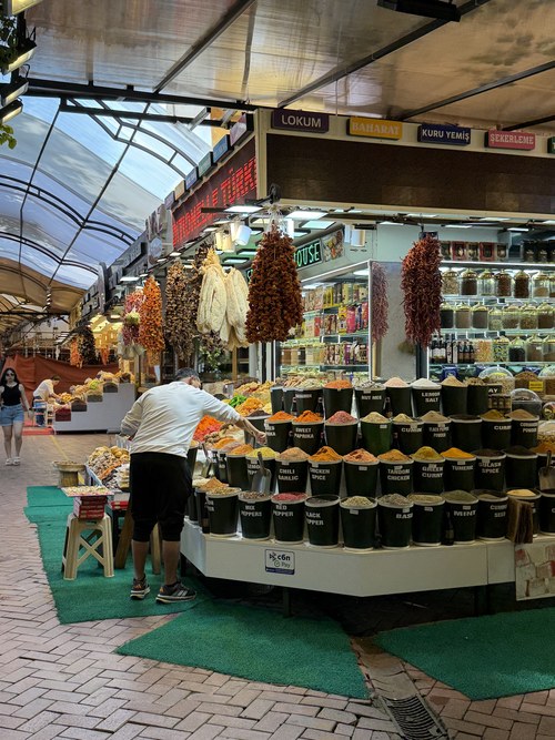 Covered market stall with buckets of spices and hanging dried peppers as a vendor works, Fethiye Old Town, Turkiye