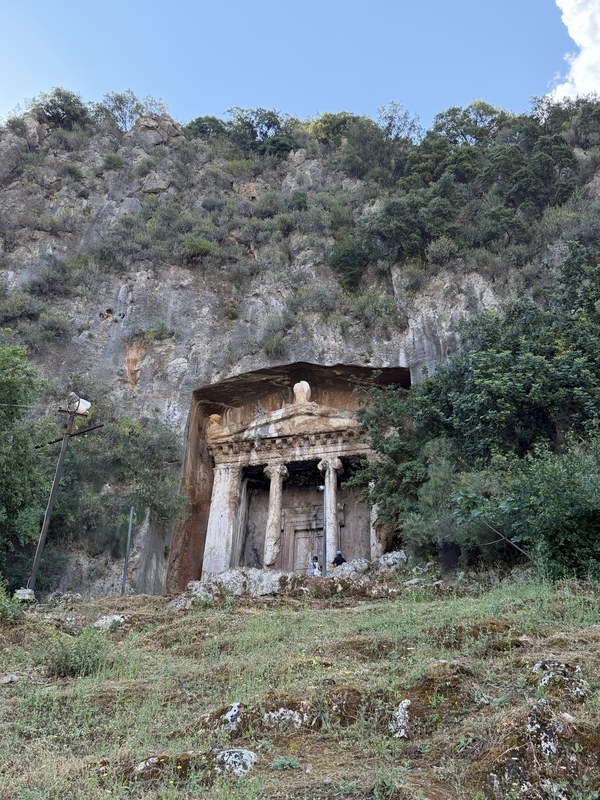 Rock-cut tomb with carved columns in a hillside cliff and a visitor standing nearby, Ancient City of Telmessos, Turkiye
