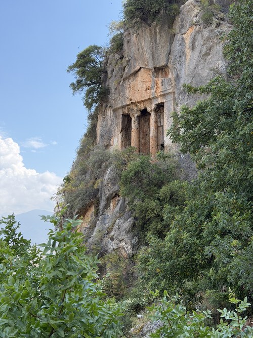 Rock-cut tomb with columned facade carved into a limestone cliff above green trees, Ancient City of Telmessos, Turkiye