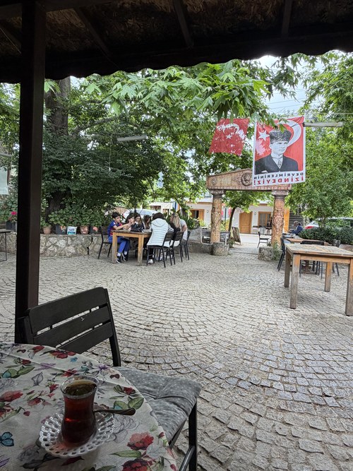 Glass of tea on a table above a shaded cobblestone village square with a group seated outside, Kuyubaşı Köy Kahvesi, Turkiye