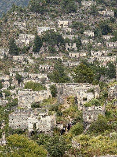 Ruined stone houses climbing a steep hillside with a small group of hikers on a dirt path, Kayaköy, Turkiye