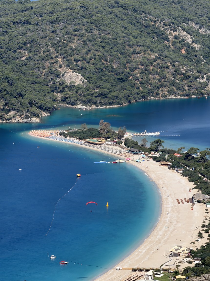 Curving sandy spit with beach umbrellas and a paraglider over the blue bay, below forested hills, Ölüdeniz, Turkiye