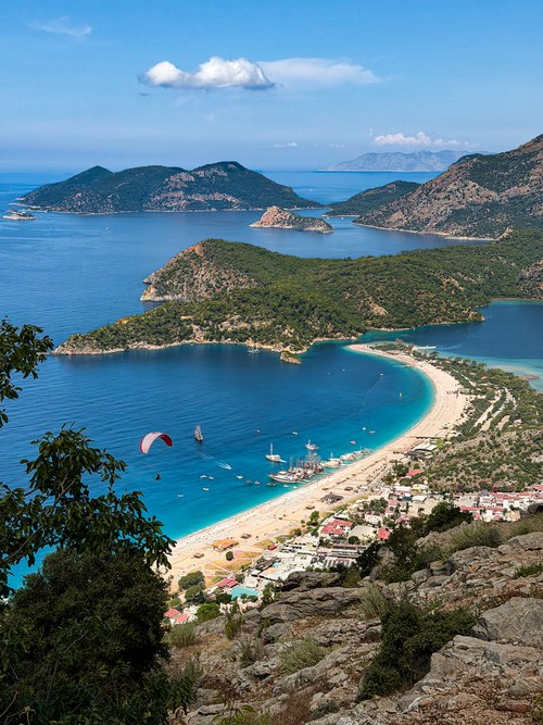 Paraglider over a blue bay with a sandy beach, tour boats and forested headlands seen from a rocky hillside, Turkiye