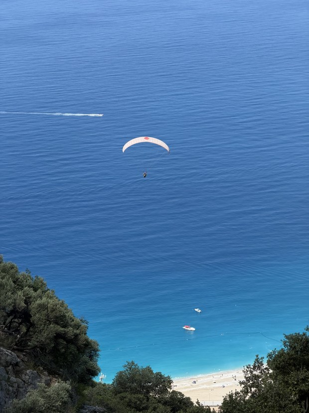Paraglider drifting over deep blue sea toward a sandy beach with small boats below, seen from a cliff, Ölüdeniz, Turkiye