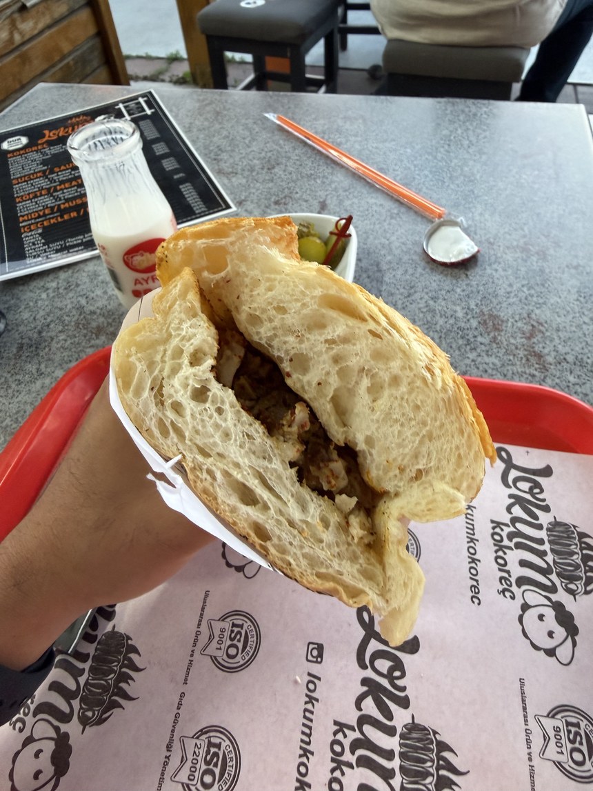 Hand holding a crusty bread sandwich with chopped meat, with ayran and pickles on the tray, Lokum Kokoreç, Turkiye