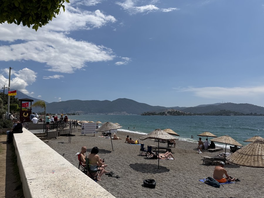 Pebble beach with straw umbrellas and sunbathers beside a lifeguard tower, with hills across the bay, Fethiye, Turkiye