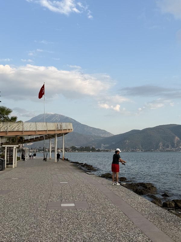 Waterfront promenade with a Turkish flag and a person fishing from the rocks, with mountains across the bay, Fethiye, Turkiye
