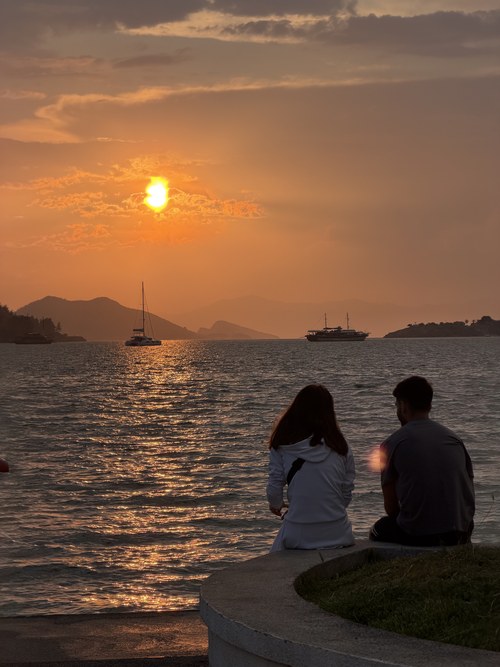 Two people sitting on a seawall watching an orange sunset over the bay with boats anchored, Fethiye, Turkiye